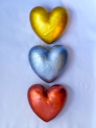Three metallic heart-shaped objects arranged vertically on a white background. The top heart is gold, the middle one is silver, and the bottom heart is bronze.