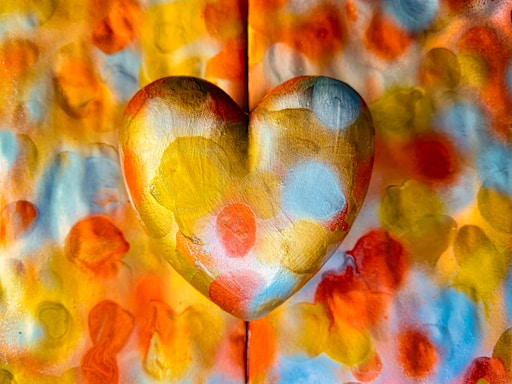 A heart shape with a golden metallic texture is prominently displayed in the center against a vibrant and colorful background. The heart and the background are covered in various bright shades of orange, red, yellow, and blue, creating a visually striking and lively composition.