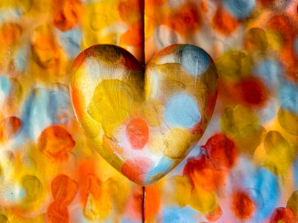 A heart shape with a golden metallic texture is prominently displayed in the center against a vibrant and colorful background. The heart and the background are covered in various bright shades of orange, red, yellow, and blue, creating a visually striking and lively composition.