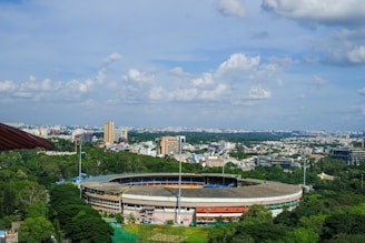 A cityscape view with a large, circular sports stadium surrounded by lush greenery in the foreground. The background features a sprawling urban skyline under a partly cloudy blue sky, with various tall buildings and a wide expanse of city structures extending to the horizon.