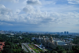 A panoramic view of a cityscape with lush greenery and a prominent, ornate government building featuring large domes. The skyline is dotted with modern high-rise buildings in the background under a partly cloudy sky.