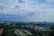 A panoramic view of a cityscape with lush greenery and a prominent, ornate government building featuring large domes. The skyline is dotted with modern high-rise buildings in the background under a partly cloudy sky.