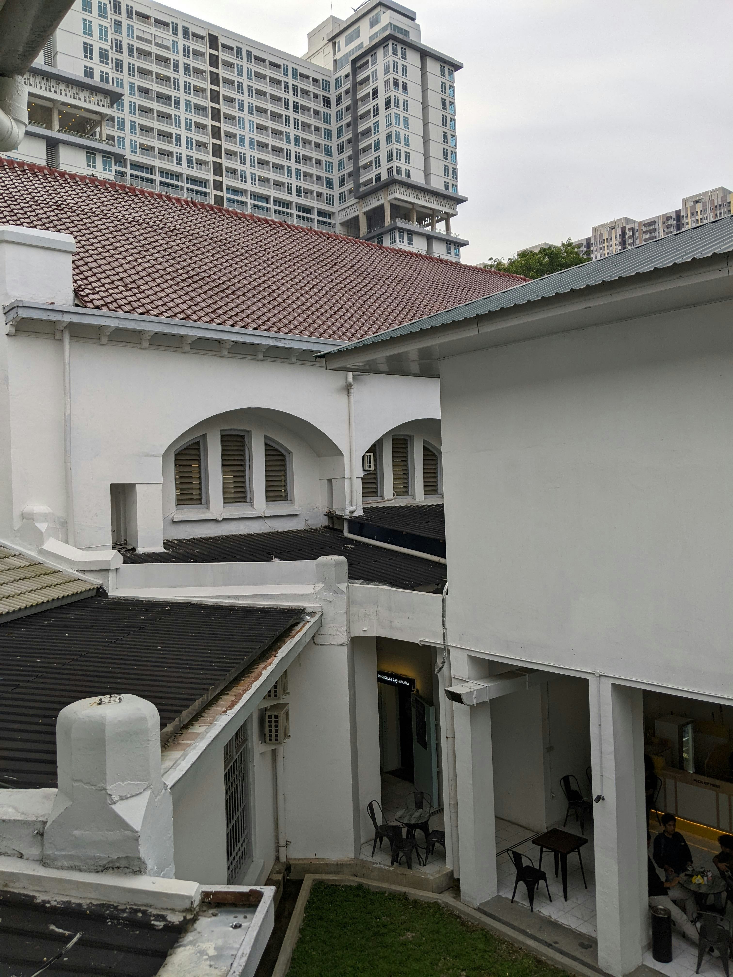 A modern high-rise building with numerous windows towers above traditional white buildings with red and gray roofs. In the foreground, the architecture features arches, and there is a small courtyard area with several empty black chairs and tables. People can be seen sitting at a table in the lower right corner, likely in an open-air indoor space.