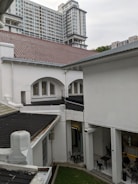 A modern high-rise building with numerous windows towers above traditional white buildings with red and gray roofs. In the foreground, the architecture features arches, and there is a small courtyard area with several empty black chairs and tables. People can be seen sitting at a table in the lower right corner, likely in an open-air indoor space.