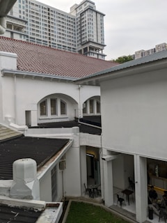A modern high-rise building with numerous windows towers above traditional white buildings with red and gray roofs. In the foreground, the architecture features arches, and there is a small courtyard area with several empty black chairs and tables. People can be seen sitting at a table in the lower right corner, likely in an open-air indoor space.