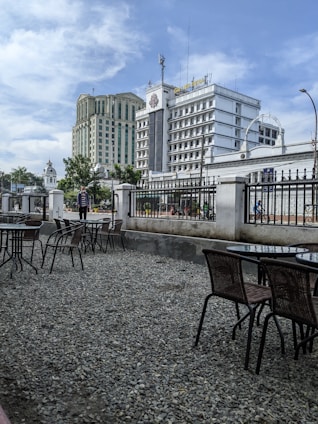 A cityscape featuring multiple high-rise buildings, including a prominent one labeled 'Grand Ménage' in the foreground. The scene includes outdoor tables and chairs placed on a gravel surface, bordered by a decorative iron fence. The sky is partly cloudy with blue tones, and there are trees and a few people visible in the background.