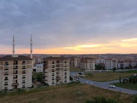 Several mid-rise apartment buildings with brown and beige exteriors sit on a grassy landscape. Two tall minarets rise next to one of the buildings. The sky is overcast with clouds, but a warm glow of sunset is visible on the horizon, casting a serene light over the urban scene. Roads curve around the buildings, with a few vehicles visible on the streets.