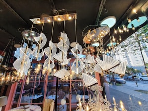 A variety of elegant and modern chandeliers are suspended from a ceiling in a showroom. The chandeliers are intricately designed with spirals and fan-like shapes, featuring a combination of metallic and crystalline elements. The lighting creates a warm, ambient glow throughout the space. Large windows reveal an outdoor street view with trees and parked cars. The interior includes decorative elements and a reflection of people seated at tables, possibly in a café setting.