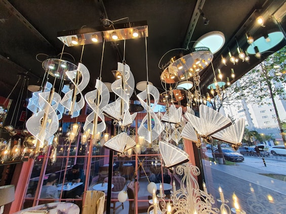 A variety of elegant and modern chandeliers are suspended from a ceiling in a showroom. The chandeliers are intricately designed with spirals and fan-like shapes, featuring a combination of metallic and crystalline elements. The lighting creates a warm, ambient glow throughout the space. Large windows reveal an outdoor street view with trees and parked cars. The interior includes decorative elements and a reflection of people seated at tables, possibly in a café setting.