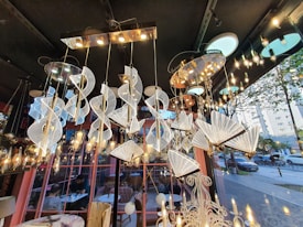 A variety of elegant and modern chandeliers are suspended from a ceiling in a showroom. The chandeliers are intricately designed with spirals and fan-like shapes, featuring a combination of metallic and crystalline elements. The lighting creates a warm, ambient glow throughout the space. Large windows reveal an outdoor street view with trees and parked cars. The interior includes decorative elements and a reflection of people seated at tables, possibly in a caf&eacute; setting.