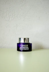 A Clinique smart clinical repair wrinkle correcting eye cream jar is placed on a light-colored surface. The jar is purple with a metallic silver lid, and the label text is in white.