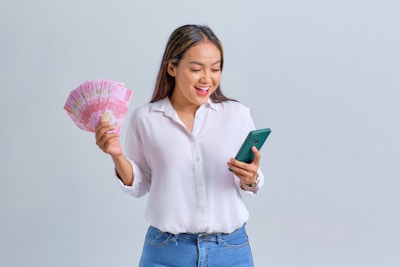 A person in a white shirt and blue jeans is holding a smartphone in one hand and several pink banknotes in the other, smiling.