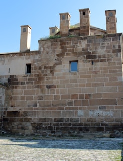 A stone wall with multiple rectangular windows is part of an old structure, featuring several chimney-like structures on the roof. The wall is constructed with irregularly shaped, large stones. The ground in front of the wall is paved with cobblestones.