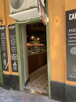 The small café entrance is inviting with menu boards showcasing various drinks and food options. Inside, there is a counter displaying pastries and other food items. The warm orange and green color scheme adds to the cozy atmosphere.