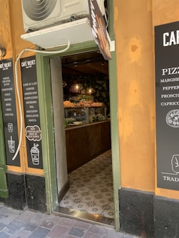 The small caf&eacute; entrance is inviting with menu boards showcasing various drinks and food options. Inside, there is a counter displaying pastries and other food items. The warm orange and green color scheme adds to the cozy atmosphere.