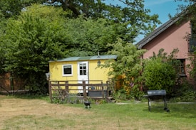 A small yellow wooden shed with a white door and window is surrounded by lush greenery and trees. In front of the shed is a wooden fence, some garden furniture, and a decorative hanging lantern. To the right, there is a larger red building partially visible, with a well-kept lawn in the foreground that includes a grill and a small fire pit.
