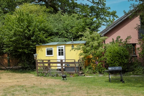 A small yellow wooden shed with a white door and window is surrounded by lush greenery and trees. In front of the shed is a wooden fence, some garden furniture, and a decorative hanging lantern. To the right, there is a larger red building partially visible, with a well-kept lawn in the foreground that includes a grill and a small fire pit.