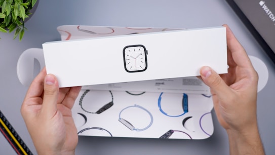 Hands holding a white box with an illustration of a smartwatch on it. The box is partially open, revealing colorful illustrations of watch bands inside. A small green plant and other packaging elements are visible in the background.