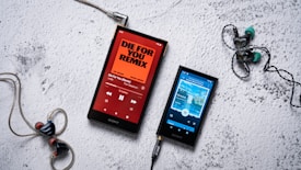 Two Sony portable music players are placed on a textured white surface. The player on the left displays an orange screen with the title 'Die for You Remix' by The Weeknd, while the player on the right shows a blue-themed screen with different music playing. The players are connected to in-ear headphones, with one black and red pair on the left and a black and turquoise pair on the right.