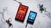 Two Sony portable music players are placed on a textured white surface. The player on the left displays an orange screen with the title 'Die for You Remix' by The Weeknd, while the player on the right shows a blue-themed screen with different music playing. The players are connected to in-ear headphones, with one black and red pair on the left and a black and turquoise pair on the right.