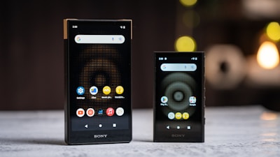 Two black Sony portable devices are displayed on a flat surface, both showing a screen with app icons such as settings, music player, and files. The background is blurred with circular light patterns.