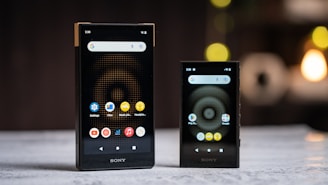 Two black Sony portable devices are displayed on a flat surface, both showing a screen with app icons such as settings, music player, and files. The background is blurred with circular light patterns.