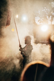 A figure stands amidst swirling smoke, holding a spear adorned with a headdress. The background is illuminated by warm, diffused lights, creating dramatic shadows and a mystical ambiance. Silhouetted trees are faintly visible, contributing to the ethereal setting.