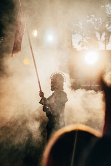 A figure stands amidst swirling smoke, holding a spear adorned with a headdress. The background is illuminated by warm, diffused lights, creating dramatic shadows and a mystical ambiance. Silhouetted trees are faintly visible, contributing to the ethereal setting.