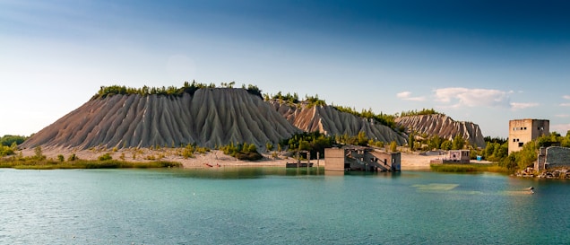 A picturesque landscape features striking, erosion-formed hills with a ridge of greenery on top. Below, a serene turquoise lake reflects the clear sky, and the foreground shows remnants of industrial buildings, creating an interesting contrast between nature and human intervention.
