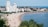 A sandy beach stretches along the coastline, populated with people enjoying the sun and water. High-rise buildings and hotels line the edge of the beach, set against a backdrop of lush greenery. The sea is calm with small waves, and swimming areas are marked with buoys. People are scattered across the beach, with umbrellas and sunbeds, indicating a lively seaside atmosphere.