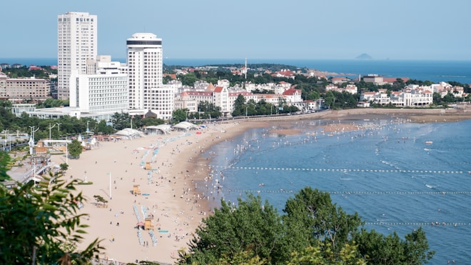 A sandy beach stretches along the coastline, populated with people enjoying the sun and water. High-rise buildings and hotels line the edge of the beach, set against a backdrop of lush greenery. The sea is calm with small waves, and swimming areas are marked with buoys. People are scattered across the beach, with umbrellas and sunbeds, indicating a lively seaside atmosphere.