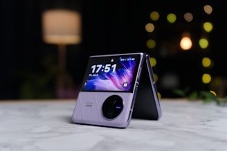 A modern foldable smartphone is placed on a marble surface in a half-open position. The screen displays the time and date with a dynamic, colorful wallpaper. The device features a sleek design with a visible camera module. In the background, there's a softly lit ambiance with blurred lights, indicating a cozy setting.