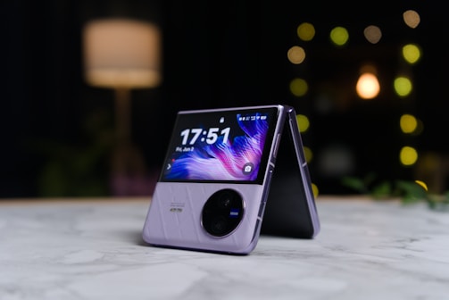 A modern foldable smartphone is placed on a marble surface in a half-open position. The screen displays the time and date with a dynamic, colorful wallpaper. The device features a sleek design with a visible camera module. In the background, there's a softly lit ambiance with blurred lights, indicating a cozy setting.