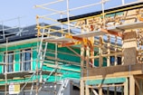 A building under construction with a wooden frame and scaffolding surrounding the structure. Green insulation material is visible on the walls, and various construction materials are present. The site signage includes safety warnings and indicates activity on the upper levels.