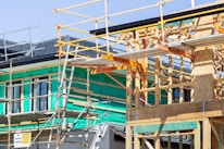 A building under construction with a wooden frame and scaffolding surrounding the structure. Green insulation material is visible on the walls, and various construction materials are present. The site signage includes safety warnings and indicates activity on the upper levels.