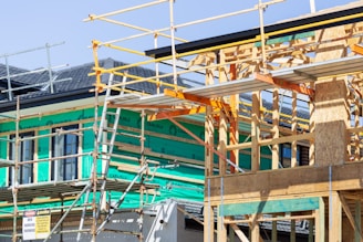A building under construction with a wooden frame and scaffolding surrounding the structure. Green insulation material is visible on the walls, and various construction materials are present. The site signage includes safety warnings and indicates activity on the upper levels.