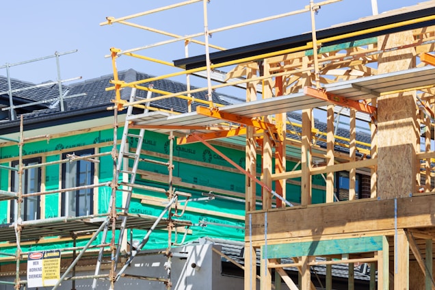 A building under construction with a wooden frame and scaffolding surrounding the structure. Green insulation material is visible on the walls, and various construction materials are present. The site signage includes safety warnings and indicates activity on the upper levels.