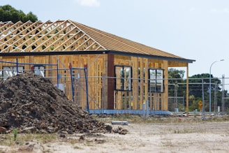 A partially constructed house with wooden framework exposed, surrounded by a temporary metal fence. There is a pile of dirt in the foreground, and the site is in a rural or suburban area with trees in the background. Construction materials are scattered around the area.