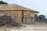 A partially constructed house with wooden framework exposed, surrounded by a temporary metal fence. There is a pile of dirt in the foreground, and the site is in a rural or suburban area with trees in the background. Construction materials are scattered around the area.