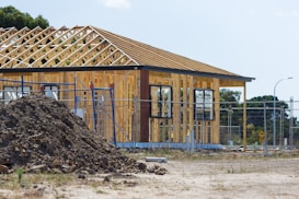 A partially constructed house with wooden framework exposed, surrounded by a temporary metal fence. There is a pile of dirt in the foreground, and the site is in a rural or suburban area with trees in the background. Construction materials are scattered around the area.