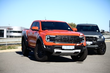 A vibrant orange pickup truck with a prominent grille stands in the foreground, accompanied by a dark gray truck in the background. Both vehicles are parked on a paved surface near a modern building under clear blue skies.