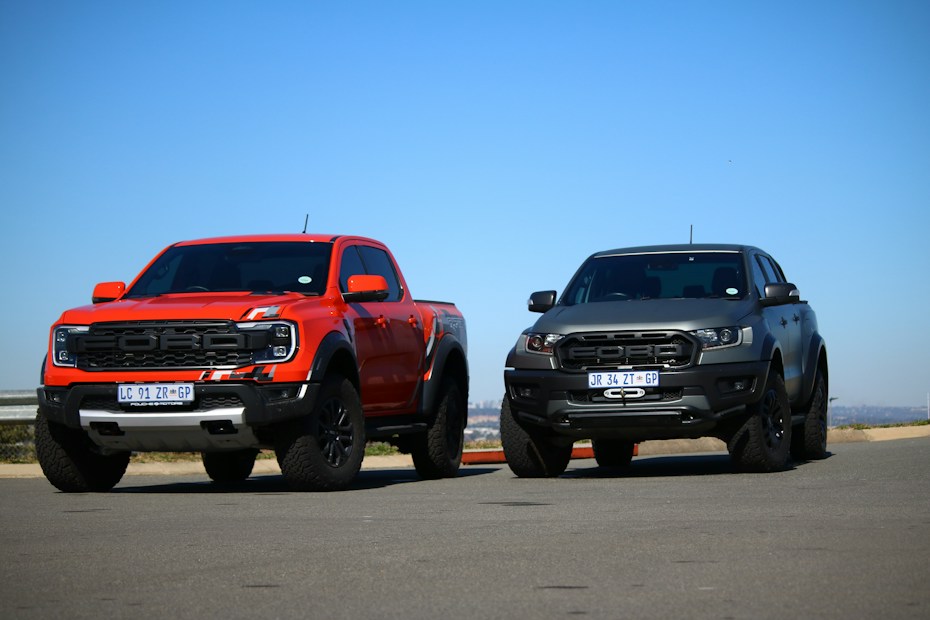 Two rugged pickup trucks are parked side by side on a paved surface. The truck on the left is painted in a vibrant red color with black accents, while the one on the right is in a matte black finish. The vehicles are positioned against a clear blue sky, conveying a sense of power and adventure.