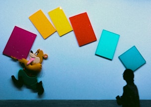 A cartoon character is joyfully running while holding a pink rectangular object in front of a wall with colorful rectangles in orange, yellow, red, and two shades of blue. A silhouetted figure stands on the right side, adding contrast.