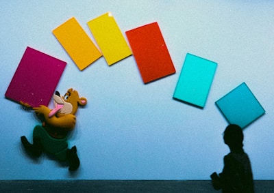 A cartoon character is joyfully running while holding a pink rectangular object in front of a wall with colorful rectangles in orange, yellow, red, and two shades of blue. A silhouetted figure stands on the right side, adding contrast.