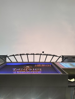 A modern architectural structure with a curved, grid-like roof design. Below is a large sign displaying text with a metallic finish. The background features a gradient sky with subtle coloration changes.
