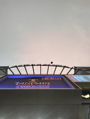 A modern architectural structure with a curved, grid-like roof design. Below is a large sign displaying text with a metallic finish. The background features a gradient sky with subtle coloration changes.