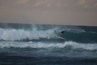 A surfer catching a massive wave under a cloudy sky.