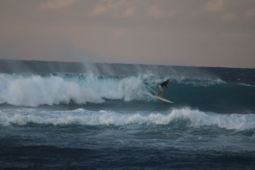 A surfer catching a massive wave under a cloudy sky.