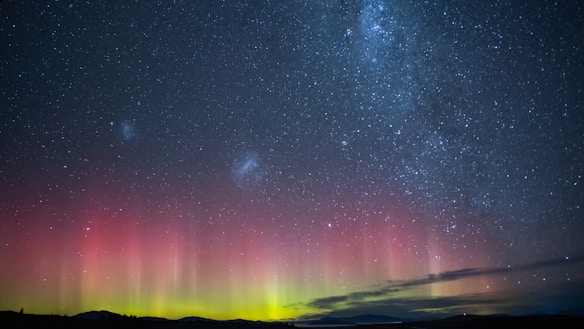 A vast night sky filled with countless stars and two prominent celestial clouds, set above a horizon lit with vibrant reds and greens resembling an aurora. The dark silhouette of a landscape can be seen at the bottom, providing contrast to the bright, colorful display above.