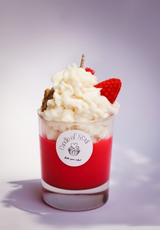 A decorative candle in a glass holder designed to resemble a dessert with red wax at the bottom and white, whipped cream-like wax swirled on top. There is a small, red strawberry-shaped detail and a stick embedded in the cream. The candle is labeled 'Candle of Soul'.